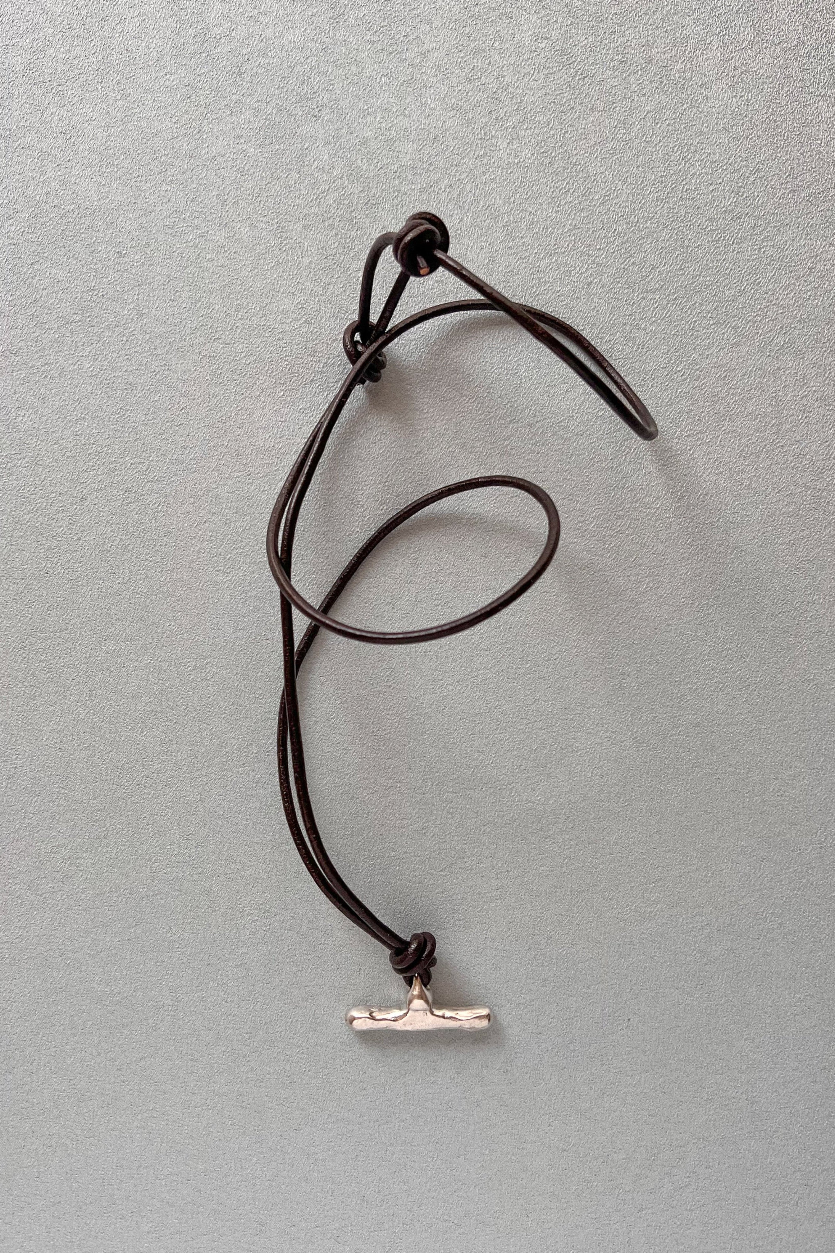 A dark brown cord necklace with a silver-toned t-bar pendant, laid out on a textured gray background.