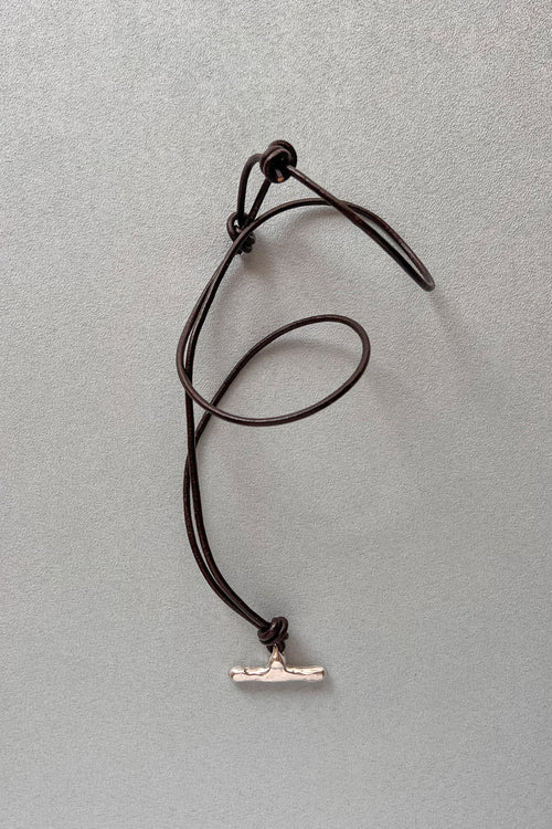 A dark brown cord necklace with a silver-toned t-bar pendant, laid out on a textured gray background.