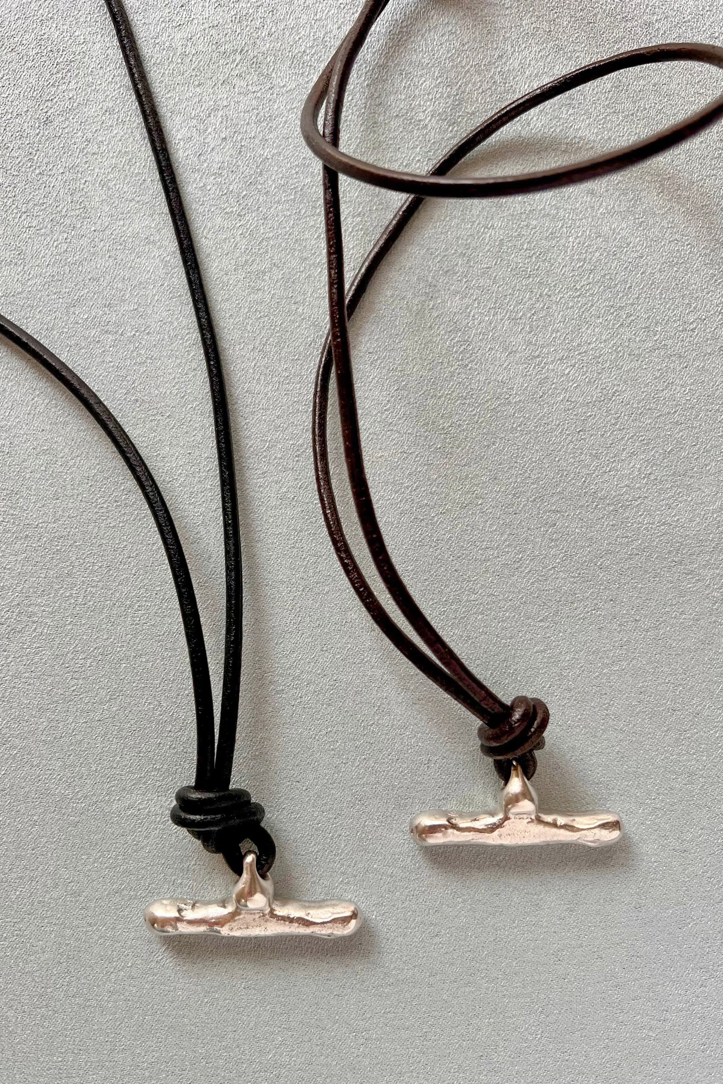 Two minimalist pendant necklaces with textured silver-toned T-shaped pendants on cords, one black and one dark brown, displayed on a light gray textured background.