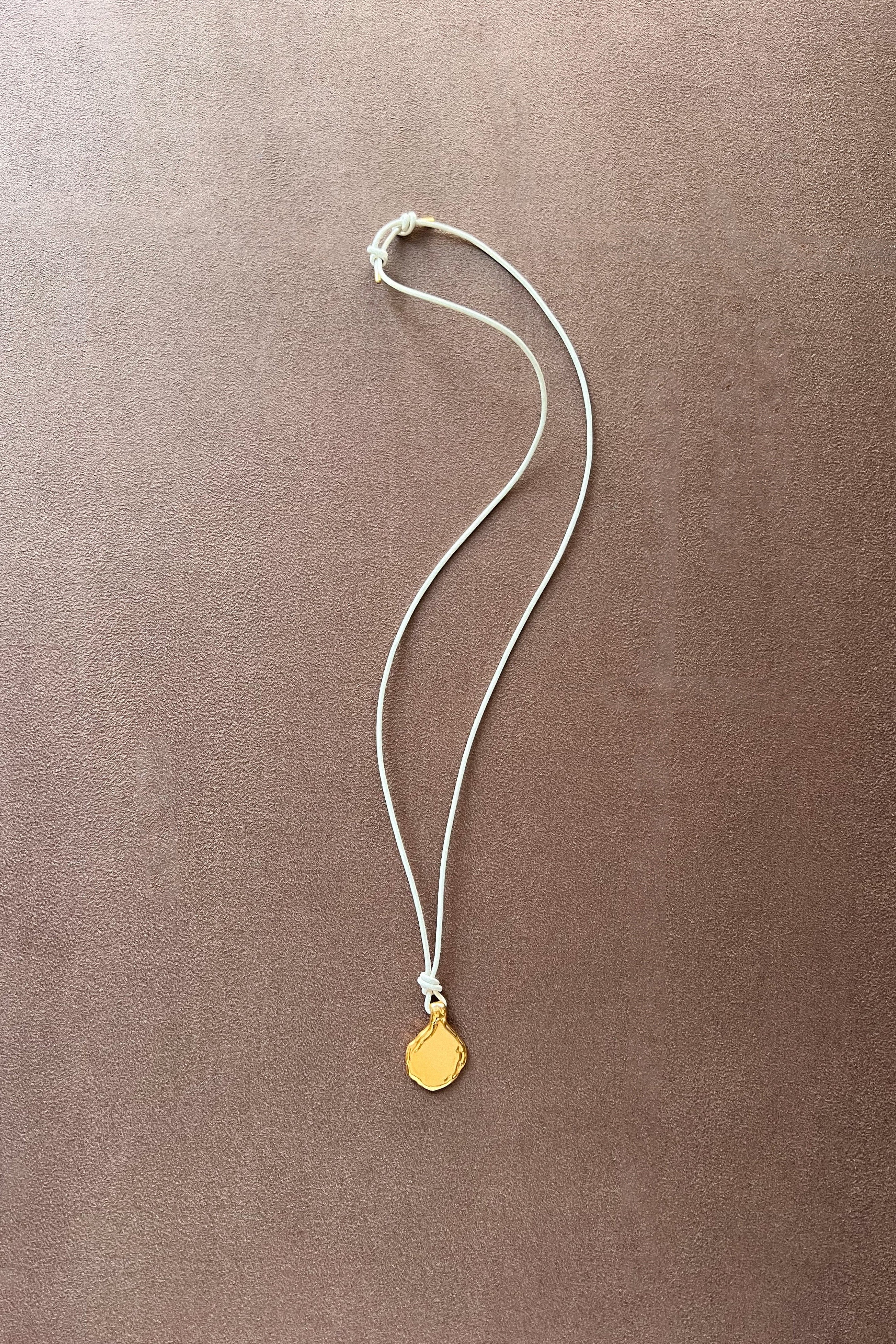 A delicate white cord necklace with a small, organic-shaped gold pendant, laid flat on a textured brown surface.