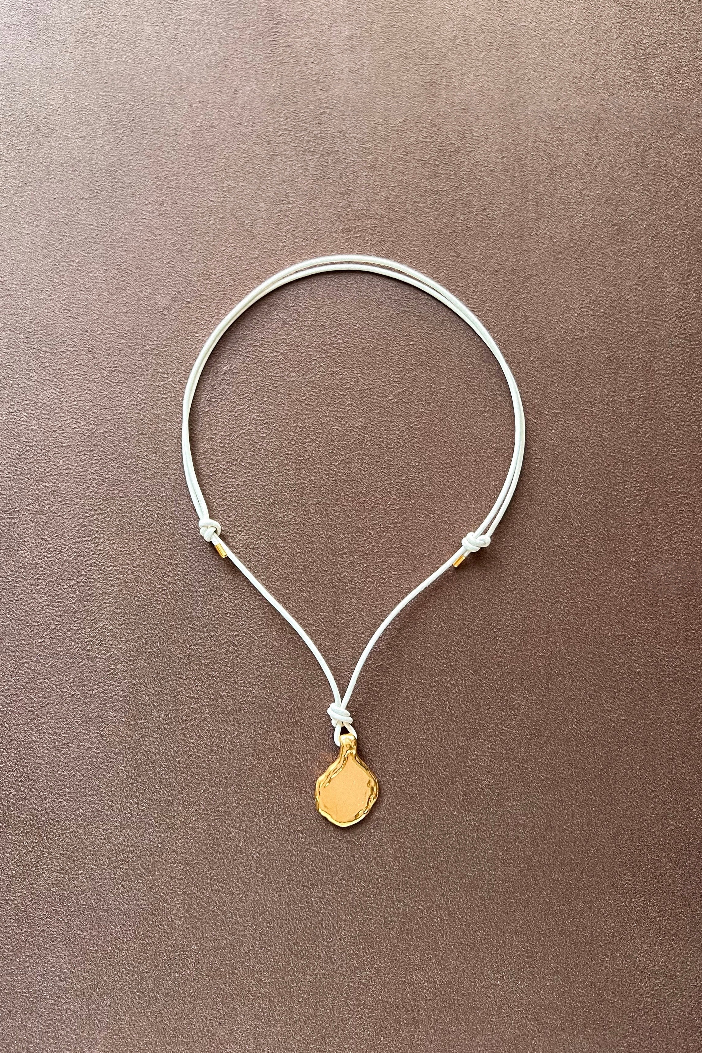 A delicate white cord necklace with a small, textured gold pendant, displayed on a textured brown background.