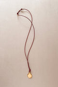 A delicate necklace featuring a small, polished gold pendant on a thin burgundy cord, laid flat on a textured beige background.